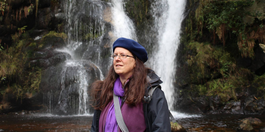 Adult female wearing purple hat stands in front of waterfall.
