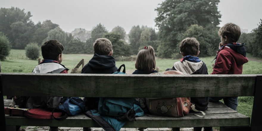Five children sit on a wooden bench looking at green trees