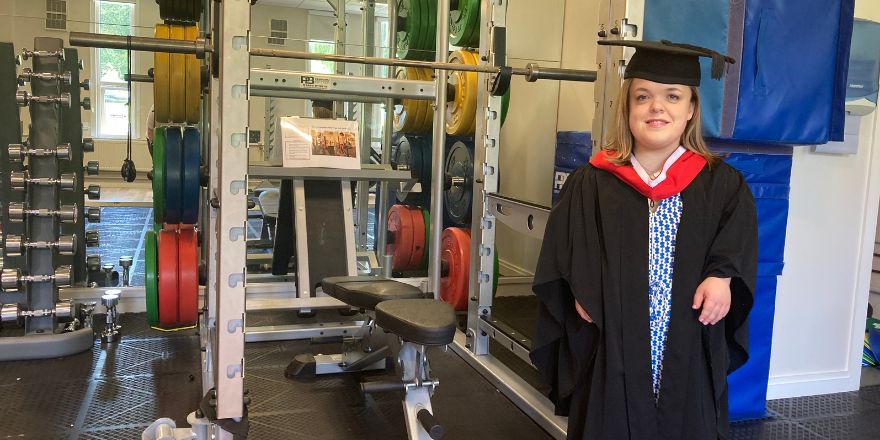 A student wearing a graduation cap and gown stands in a gym