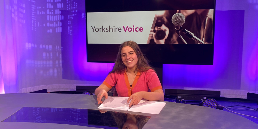 Female student sits at news desk smiling
