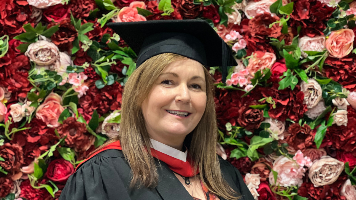 A mature female student posing against a flower wall in graduation cap and gown.
