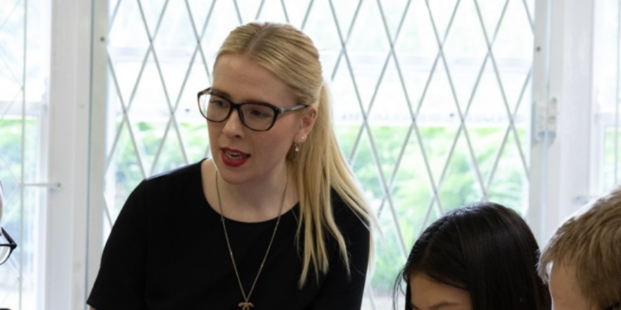 Blonde female in glasses with hair tied back and black top on speaks to students.