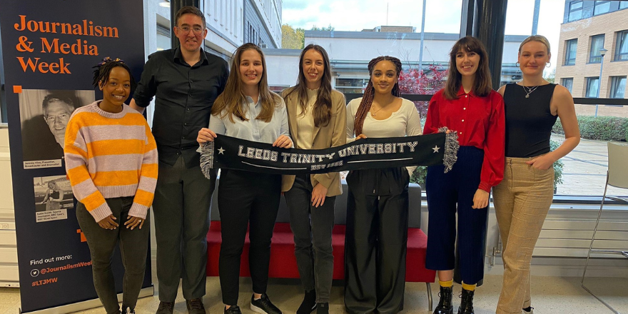 Group of students line up in front of window holding Leeds Trinity University black scarf.