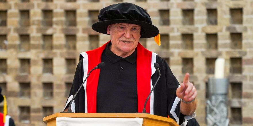Elderly male wearing cap and gown stands at lectern.