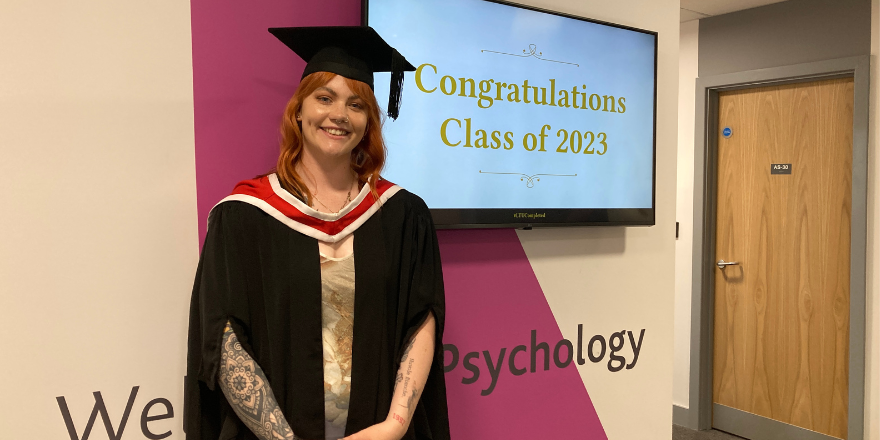 A girl with ginger hair wearing a black cap and gown poses in front of a screen that says, Congratulations Class of 2023