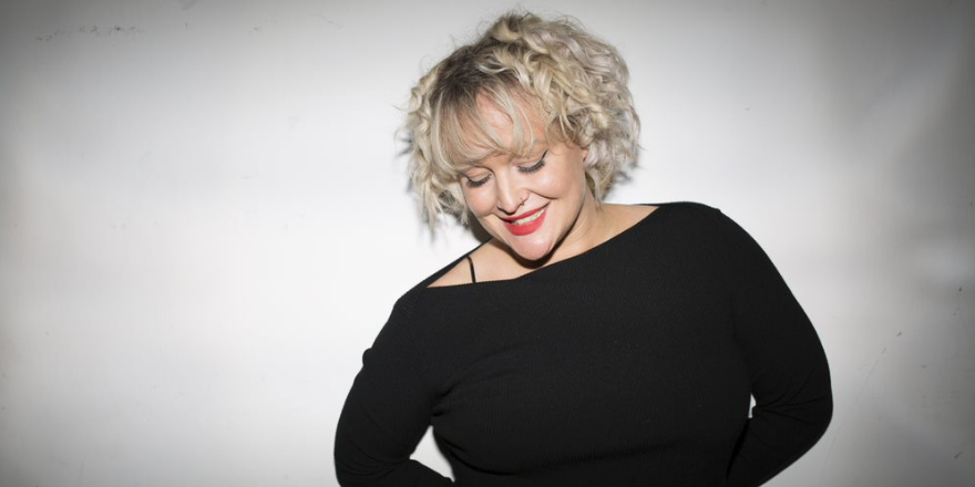 Woman with blonde curly short hair stands against white background wearing black off the shoulder long sleeved top.