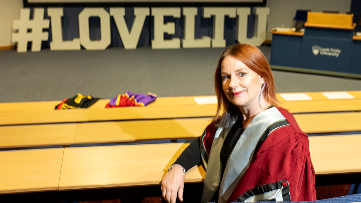 An academic in a red gown sitting in a lecture theatre in front of a sign that reads Love LTU.