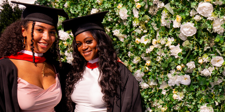 Two female Leeds Trinity graduates stand side by side with graduation caps on in front of a flower wall.