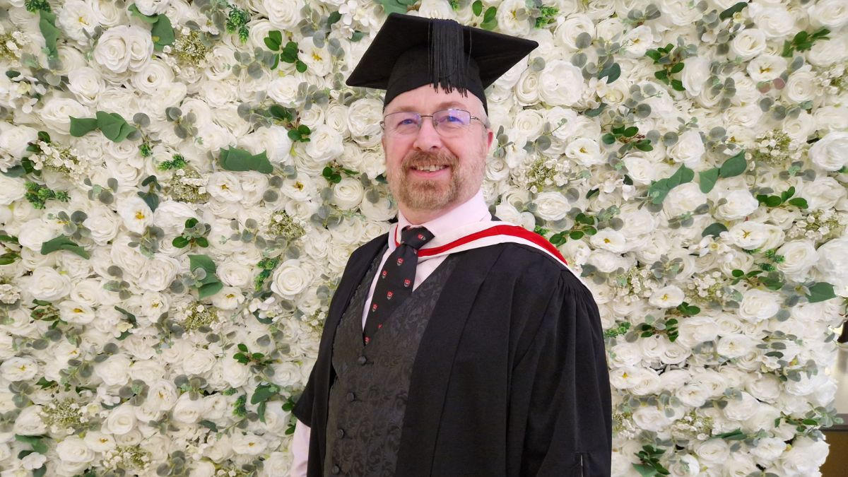 A graduate wearing a cap and gown stands in front of a white flower wall