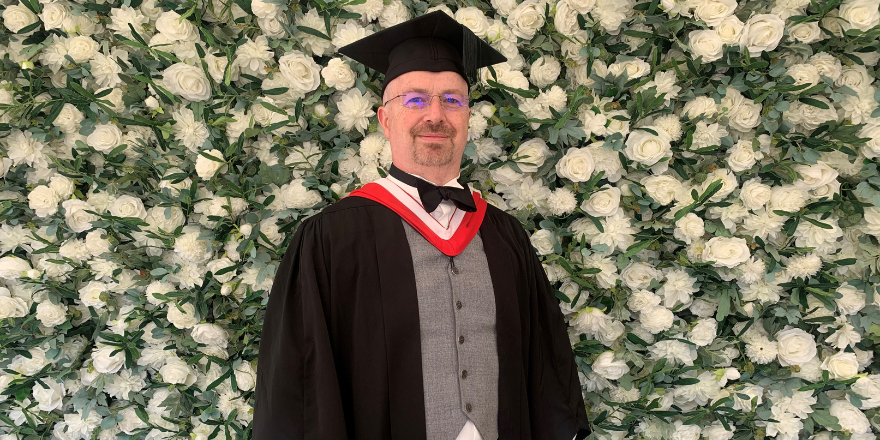 Male graduate stands in front of flower wall wearing graduation cap and gown.