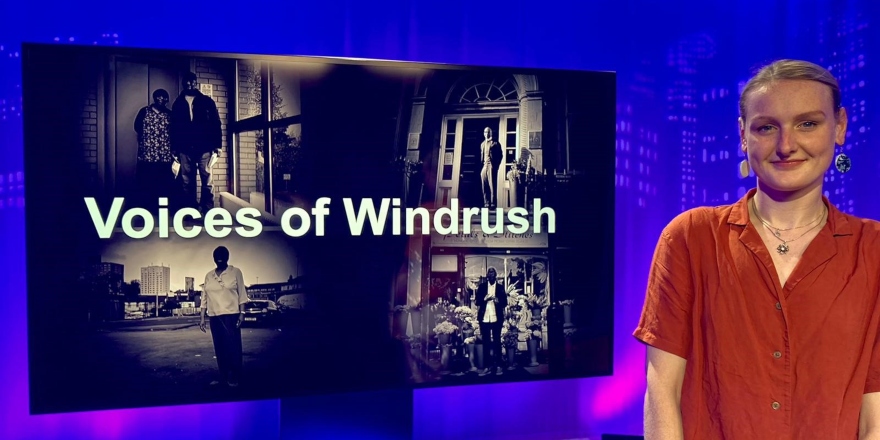 Blonde student in red blouse next to black and white TV screen which reads 'Voices of Windrush'.