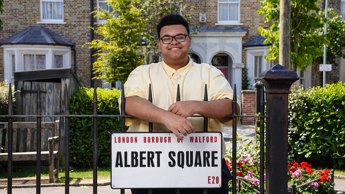 A Leeds Trinity University alumnus wearing a pale yellow shirt and smiling and posing behind a low iron fence with a white sign reading Albert Square on it.
