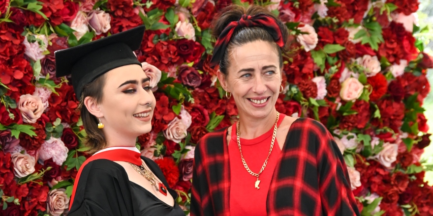 Brunette female graduate in black graduation cap and gown with brunette woman in red top and checked jacket.