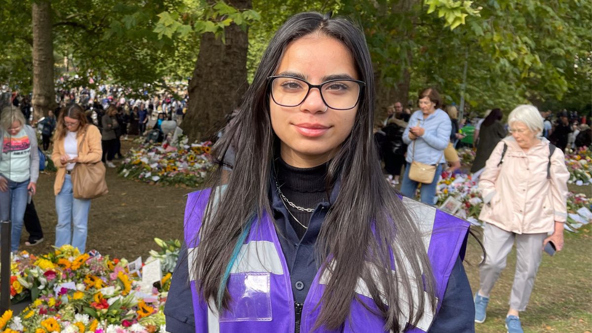 A girl with dark hair and glasses in a purple vest smiles for a picture in front of trees and flowers