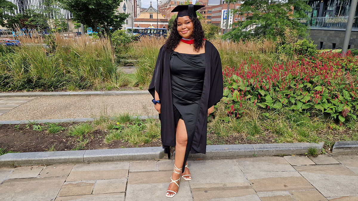 A student in graduation cap and gown smiling in a green space in Leeds.
