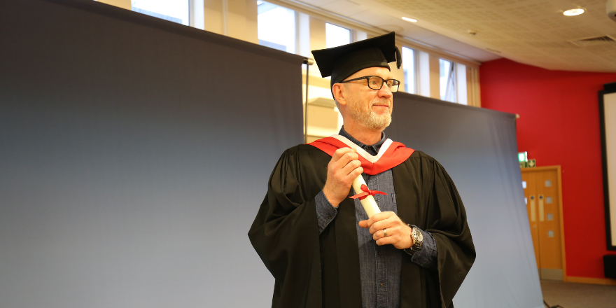 Mature male student in glasses and graduation cap poses for photo.