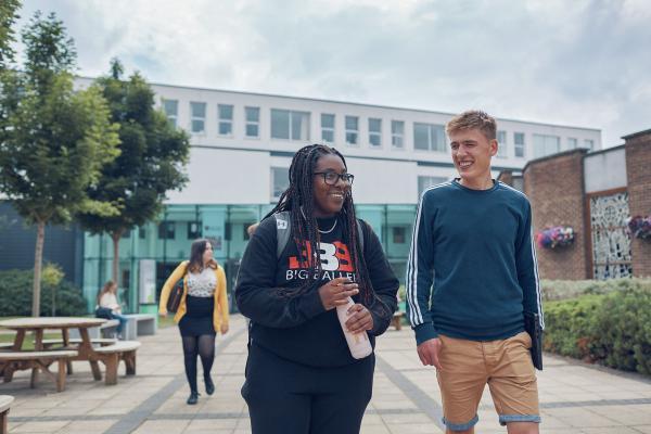 Two university students walk together and chat on a paved campus path, smiling and relaxed. Behind them, two other students are walking in the same direction. The scene is set outside a modern academic building with trees, benches, and flower displays.