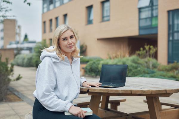 A student sits at a round wooden picnic table on a university campus, with a laptop in front of her and a notebook in hand. She is wearing a light grey hoodie and smiling at the camera. Modern student accommodation buildings and landscaped greenery are visible in the background.